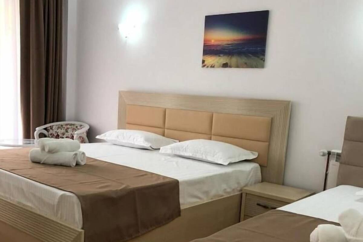 A hotel room with two beds, a wooden floor, a painting on the wall, and a chair with a floral cushion.