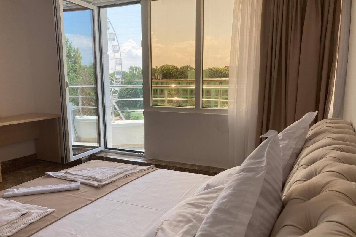 A bedroom with a bed, a window with curtains, and a view of greenery and buildings outside.
