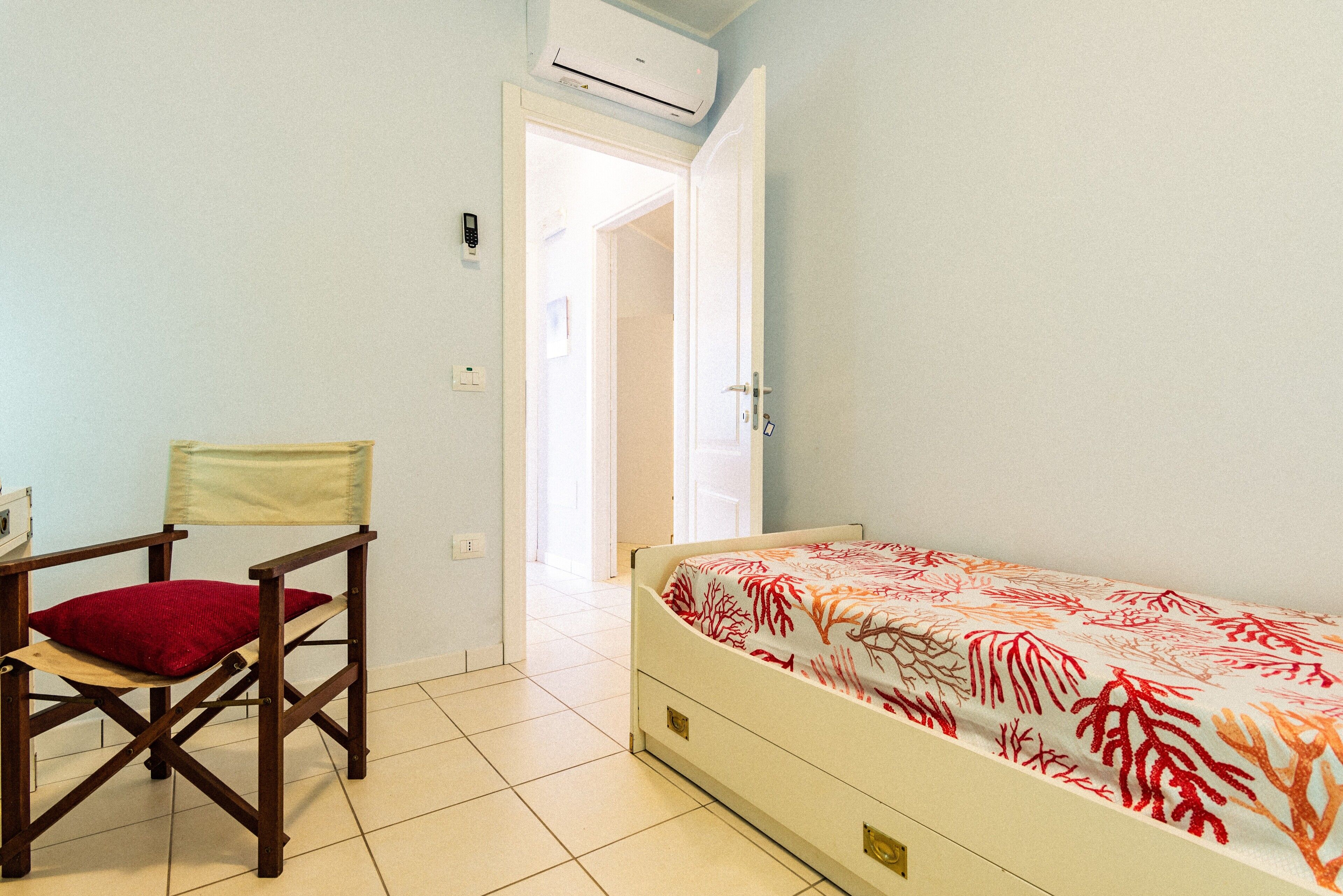 A room with a bed, a chair, an air conditioning unit, and a door leading to another room.