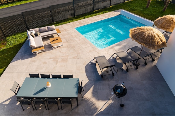 Un patio trasero con piscina, una zona de estar con sofá y sillones, una mesa de comedor con sillas y dos sombrillas tiki.