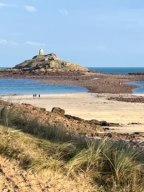 Eine Küstenlandschaft mit einer Felsformation, einem kleinen Gebäude darauf und einem Sandstrand mit einigen Personen.