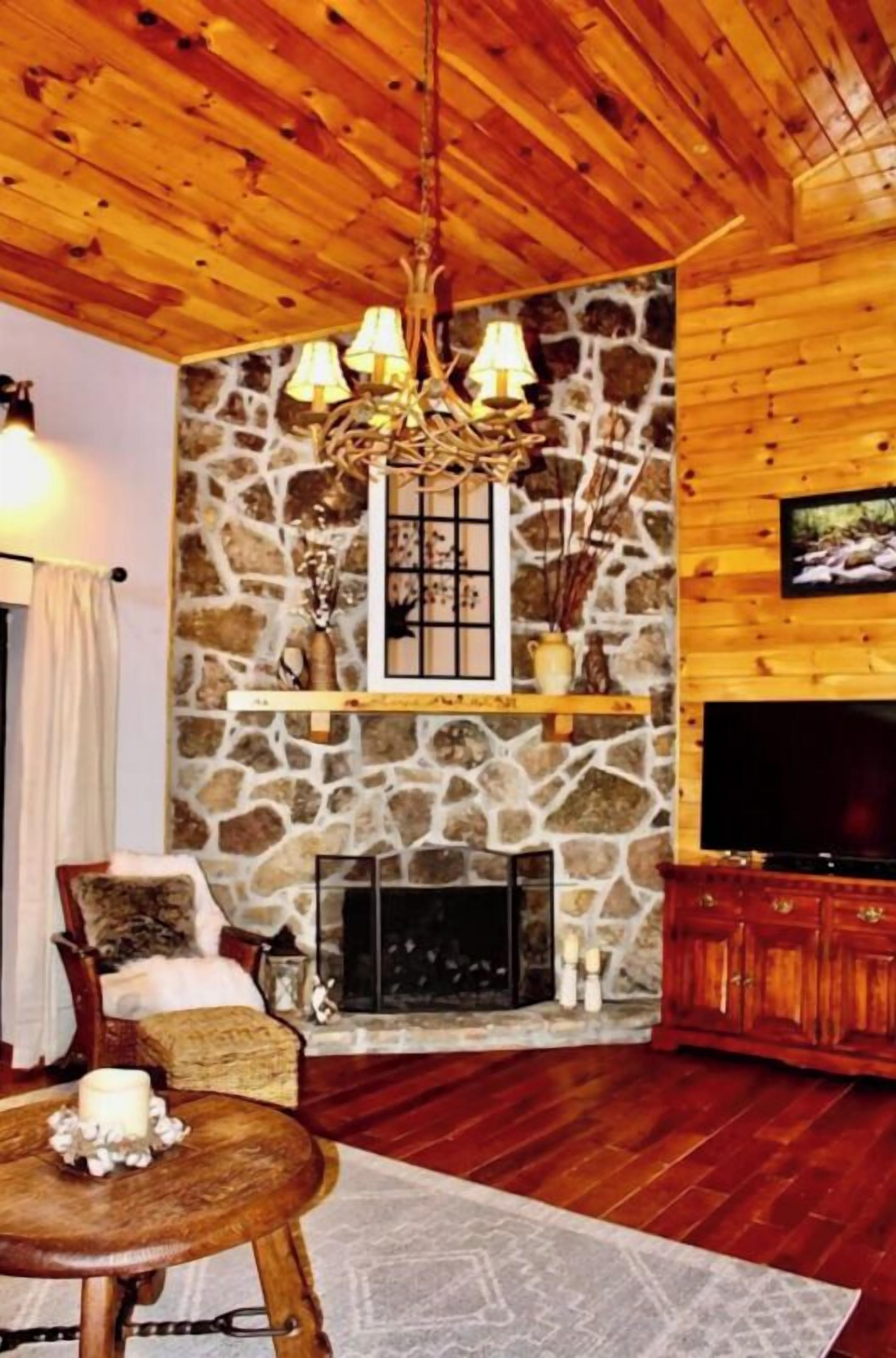 A cozy living room with wooden walls, a stone fireplace, a television, and a chandelier.