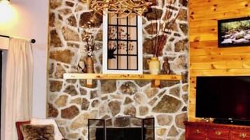 A cozy living room with wooden walls, a stone fireplace, a television, and a chandelier.