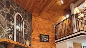 Un salon chaleureux avec une cheminée en pierre, des murs en bois, un téléviseur et un balcon doté d’une balustrade en fer forgé.