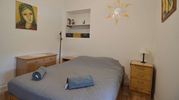 A bedroom with a bed, a nightstand, a lamp, a wall shelf with books, and a painting. - Appartement Proche de la Plage - Wifi, Bien Agencé (Asnelles)