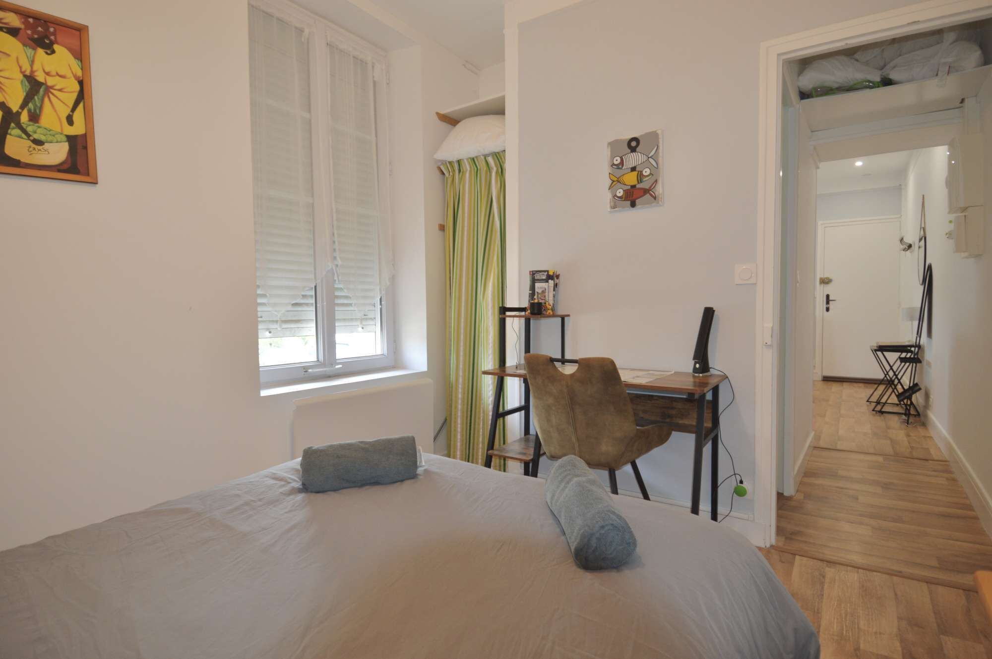 A bedroom with a bed, a desk, and a chair. There is a window with blinds, a curtain, and a painting on the wall.