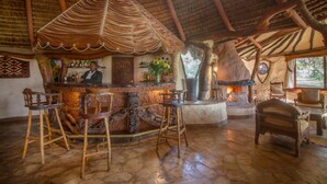A rustic interior with a bar area, wooden chairs, a fireplace, and a stone floor. - Chui Lodge (Naivasha)