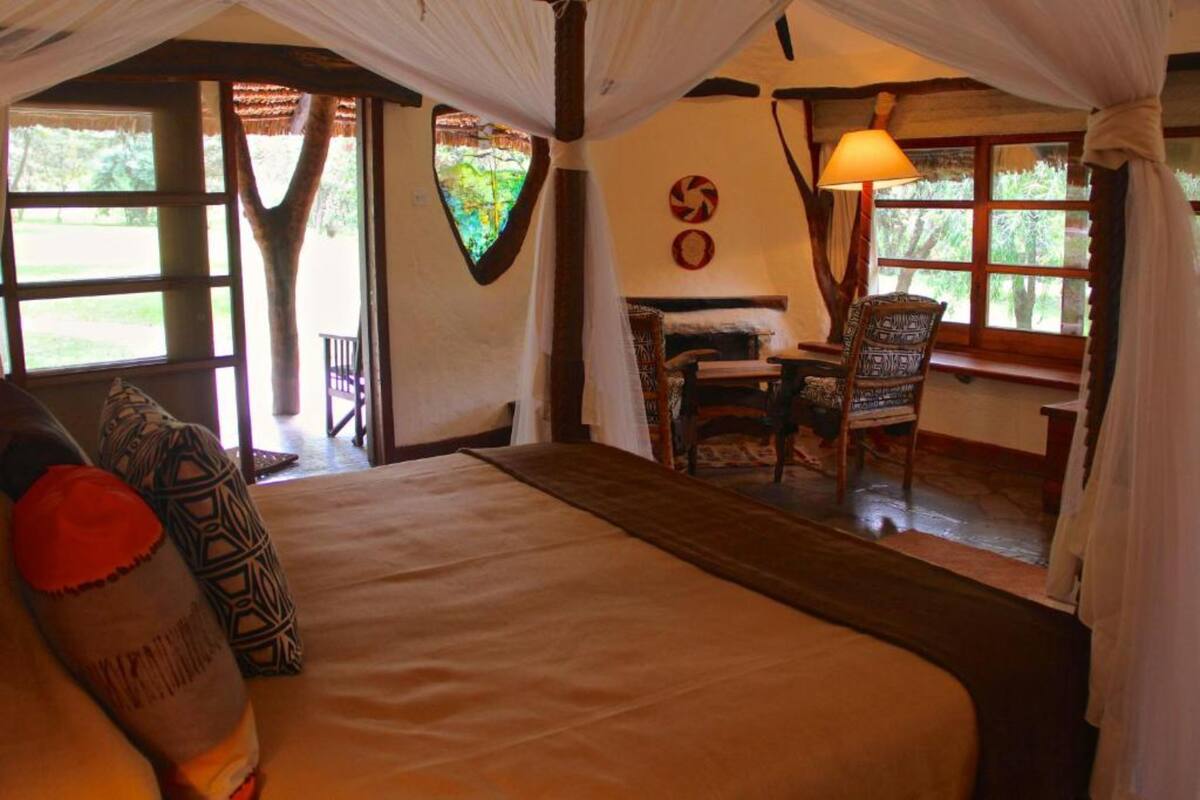 Um quarto aconchegante com cama, mesa e uma cadeira. Há uma janela com cortinas e um abajur.