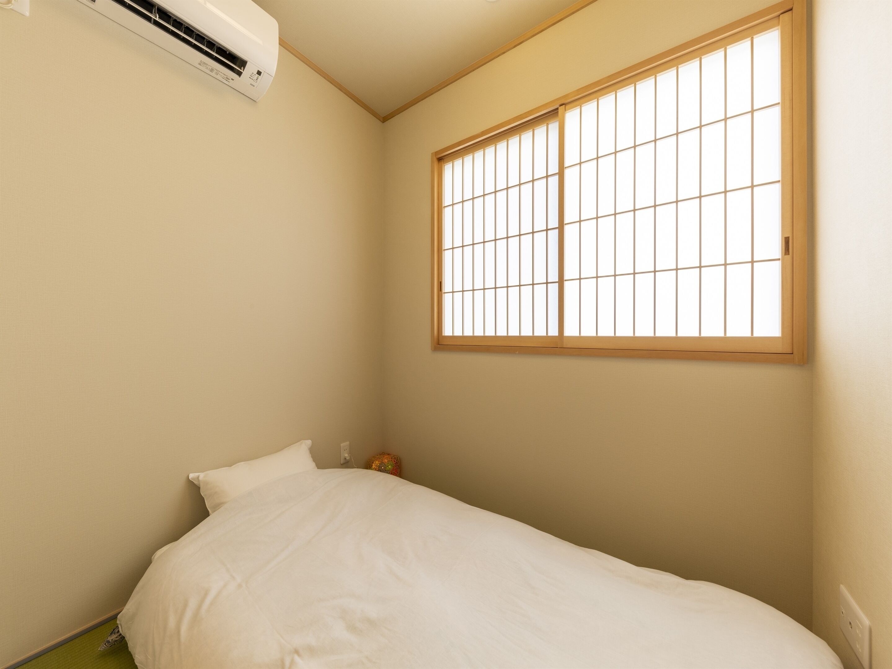 A small, single-bed room with a window, an air conditioning unit, and a bedside table.
