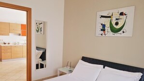 Un cuarto de hotel con una cama, una mesita de noche, un cuadro y vista a la cocina.