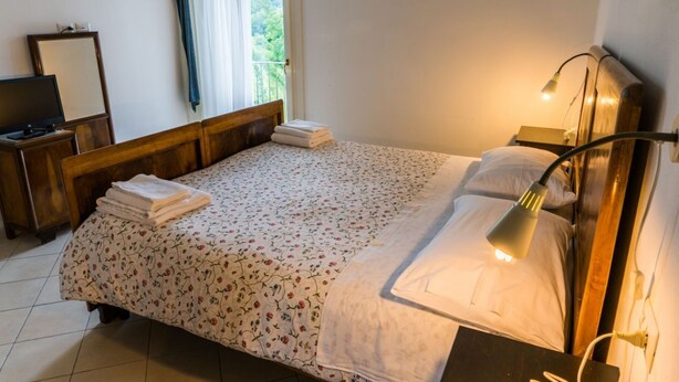 Un lit bien fait, recouvert d’une couvre-lit à motifs floraux et de draps blancs, une lampe de chevet et une vue sur la verdure par la fenêtre.