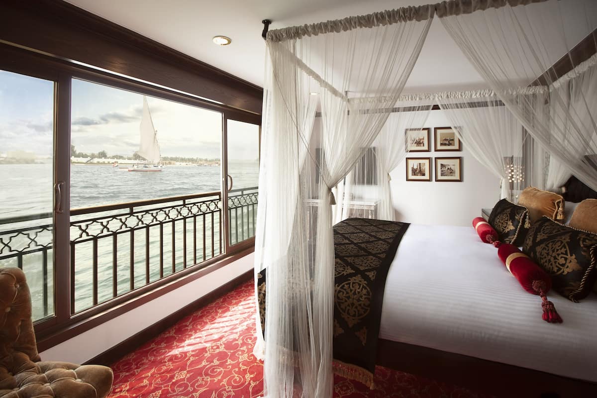 A bedroom with a large window, a bed with red and gold pillows, and a canopy.