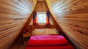 Una habitación pequeña y acogedora con paredes de madera y techo inclinado, que presenta una cama con colcha y almohadas rojas, una mesita de noche con lámpara, y una ventana con cortinas.