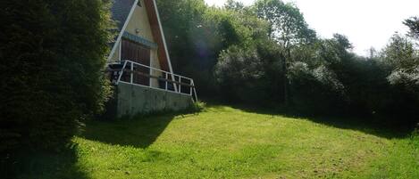 A small, triangular wooden structure with a balcony, surrounded by a grassy area and dense trees.