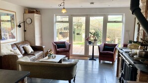 Ein gemütliches Wohnzimmer mit einer braunen Ledersofa, einem weißen Sessel und einem Couchtisch. Ein großes Fenster bietet Blick auf den Garten.