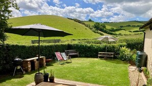 Ein Garten mit einer Holzterrasse, einem Grill, einem Picknicktisch und einem großen Sonnenschirm.