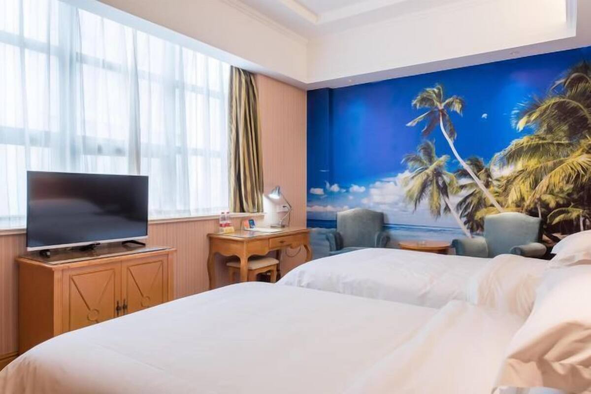 A hotel room with a bed, a desk with a computer, a TV, and a mural of palm trees and a beach.