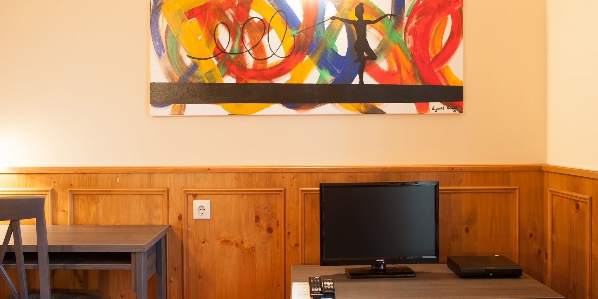 A room with a wooden paneled wall, a flat-screen TV, a laptop, and a remote control on a wooden table.