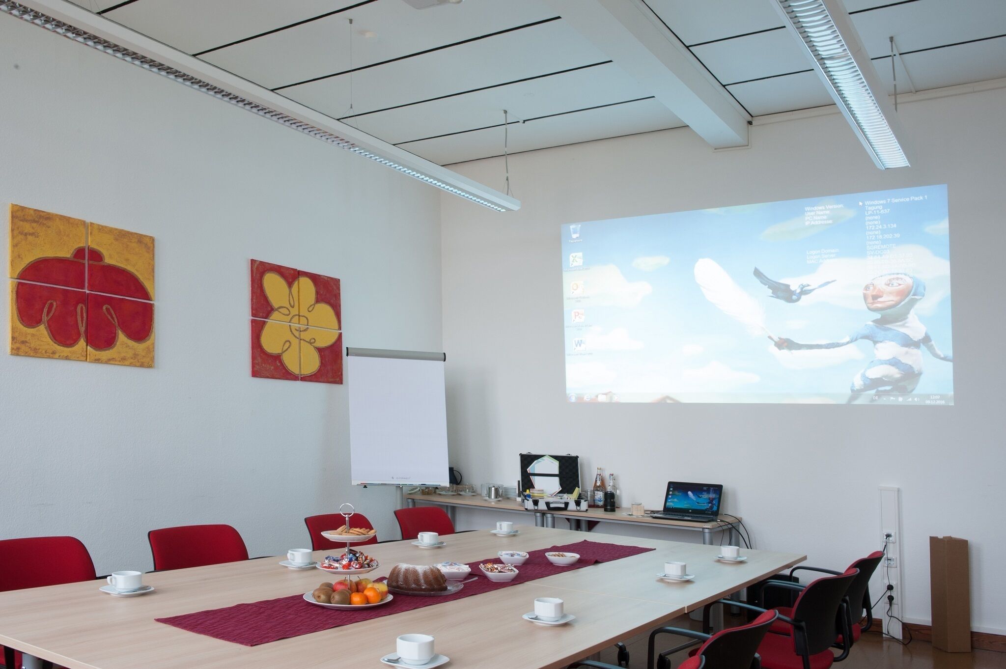 A conference room with a long table, red chairs, a projector screen, and two flower paintings on the wall.