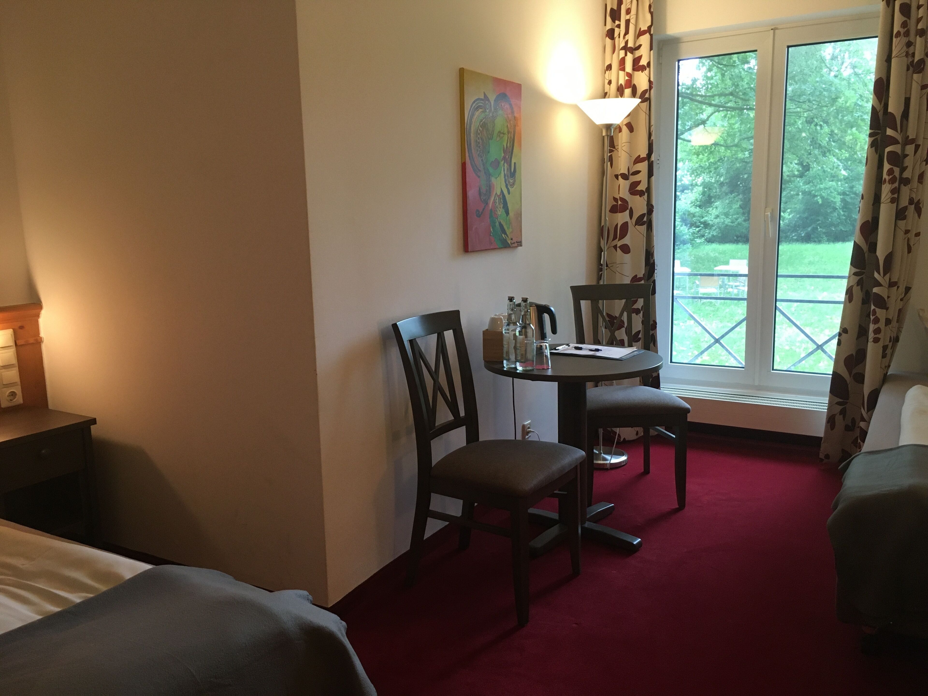 A hotel room with a bed, a desk with a lamp, a chair, a small table with a lamp, and a window with a view of greenery.