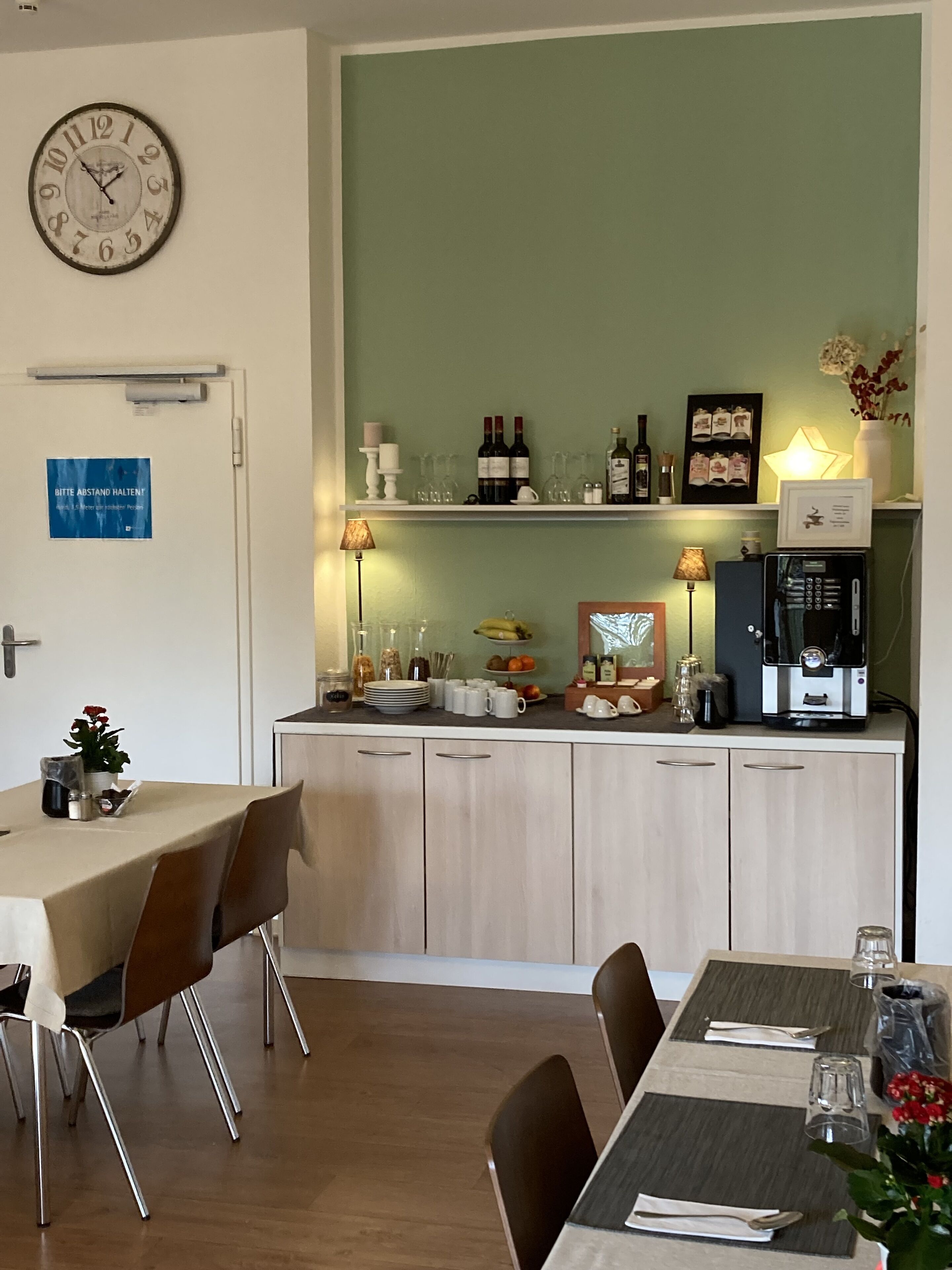 Un comedor con una mesa dispuesta para dos personas, un reloj en la pared y una zona de cocina con una cafetera y botellas de vino.