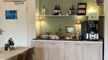 Un comedor con una mesa dispuesta para dos personas, un reloj en la pared y una zona de cocina con una cafetera y botellas de vino.