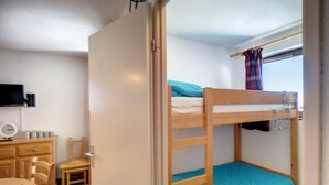 Een stapelbed met een blauwe matras, een houten bureau met stoel, een klein tafeltje en een televisie aan de muur.