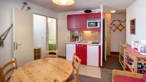 Een compacte keuken met rode kastjes, een magnetron en een vaatwasser. Er is een houten eettafel met stoelen, en een klein kitchenette gedeelte met een koffiezetapparaat en een plank met diverse spullen.