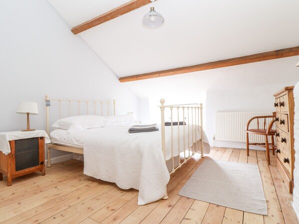 A bedroom with a wooden floor, a bed with white bedding, a nightstand with a lamp, a dresser, and a radiator. - Trelash Cottage (Launceston)