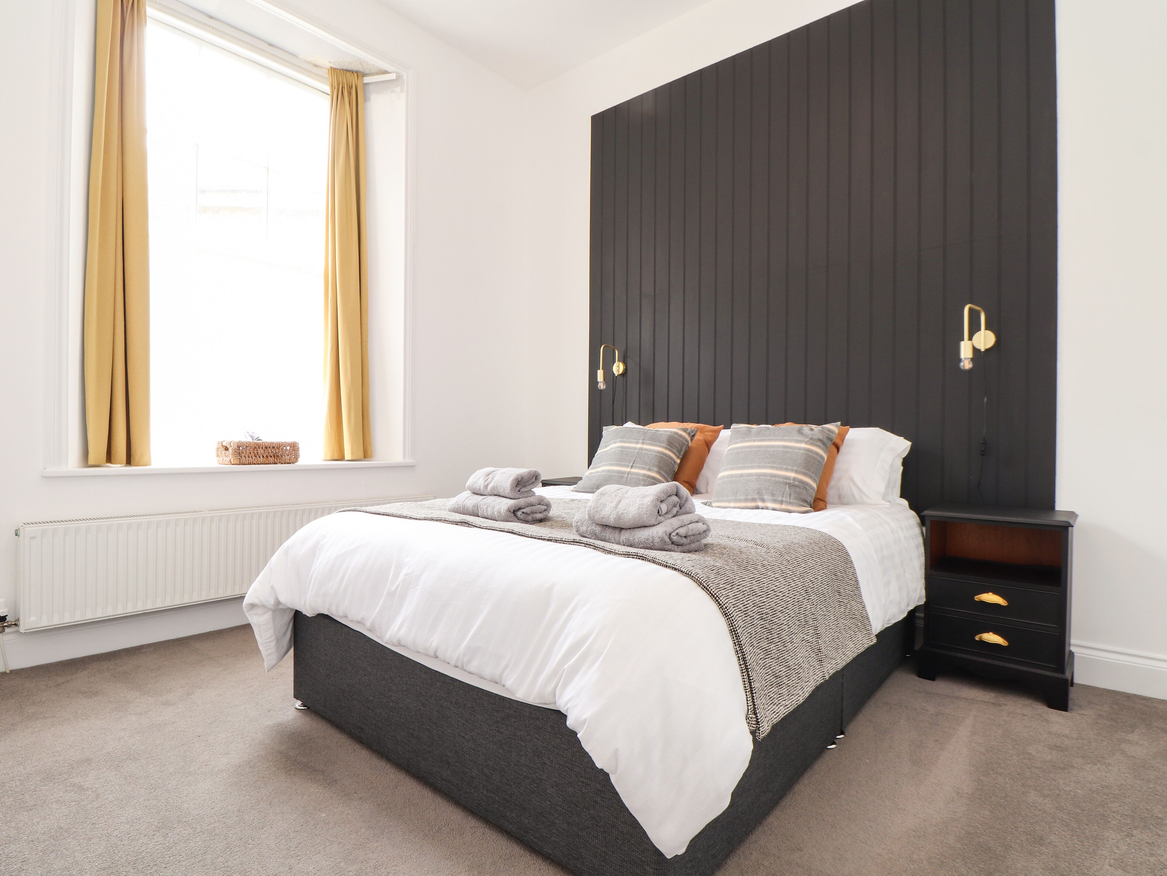 A bedroom with a bed, a bedside table, a nightstand, a window with curtains, and a radiator.