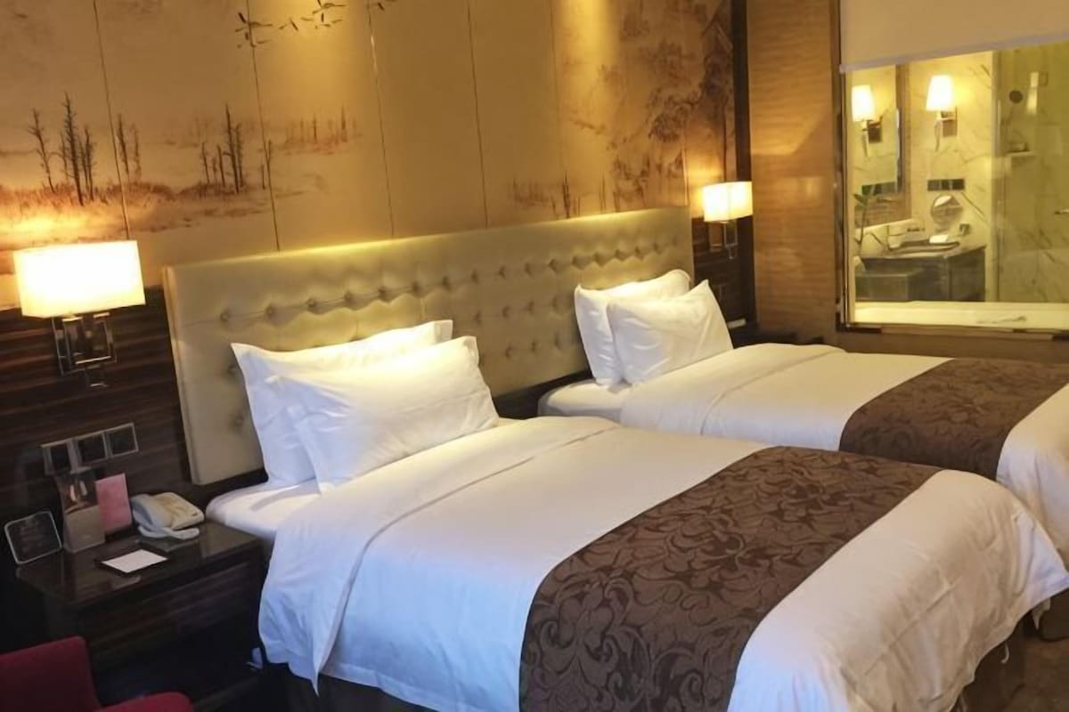 A hotel room with two beds, a nightstand with a phone, and a bathroom visible through a glass door.