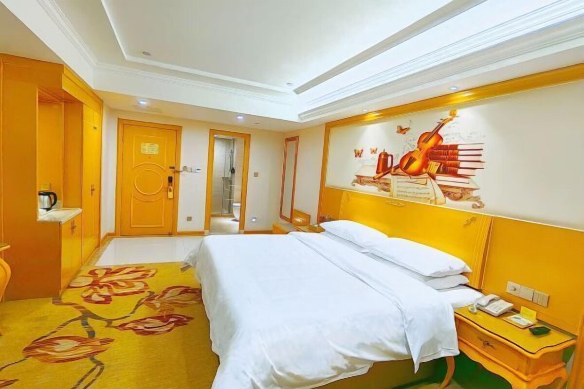 A hotel room with a bed, a bedside table with a phone, a wall with a musical instrument and books, and a door leading to another room.
