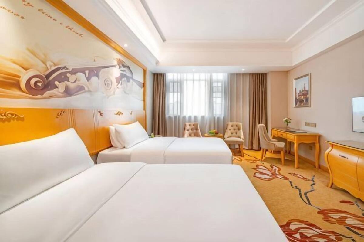 A hotel room with two beds, a desk, a chair, a television, and a large window with curtains.