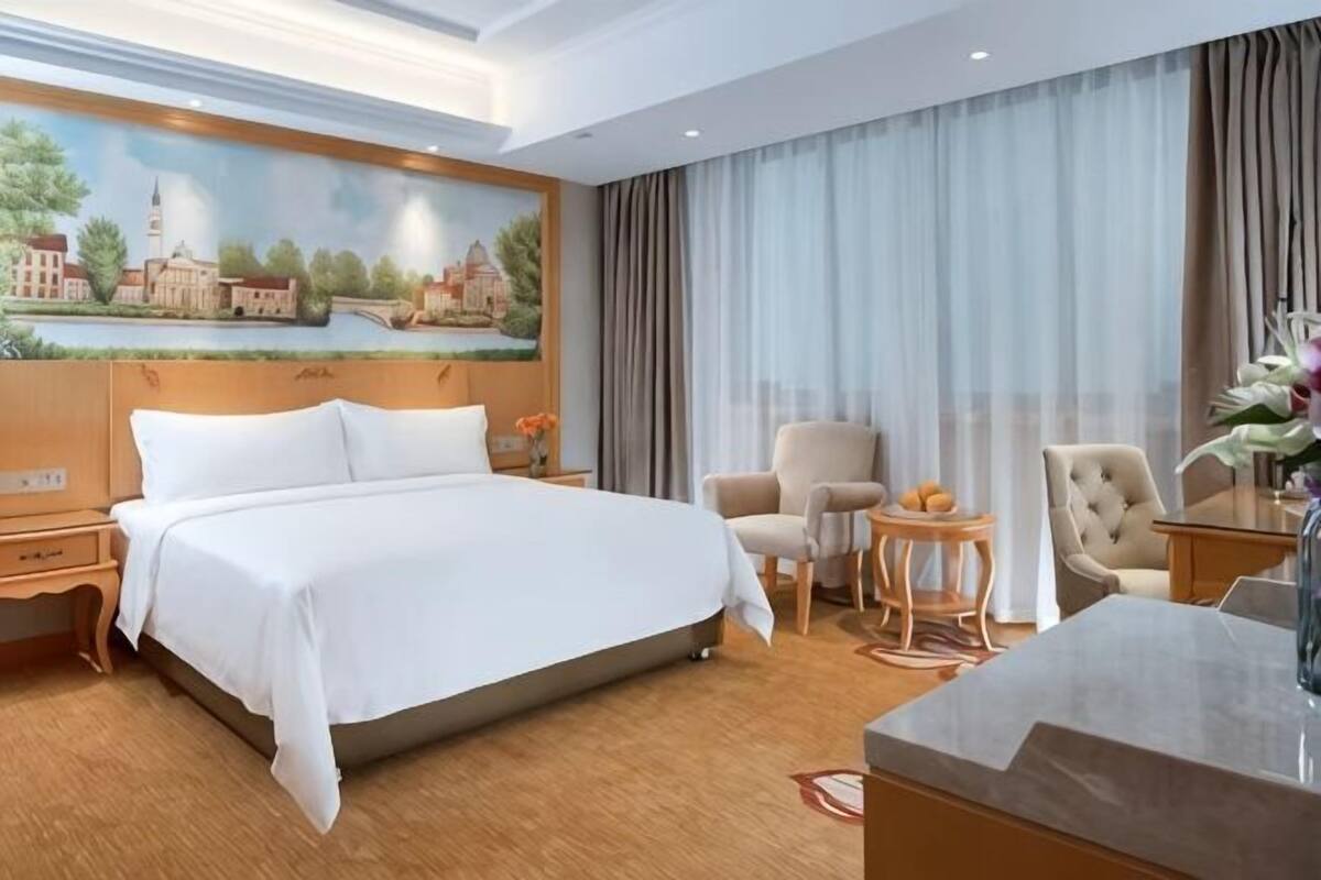 A hotel room with a large bed, two armchairs, a small round table, a desk, and a painting on the wall.