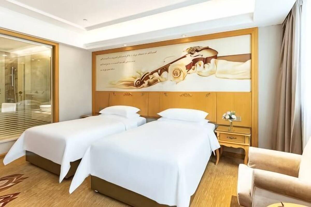 A hotel room with two beds, a wooden headboard, a large floral artwork, a wooden nightstand, and a view of a glass shower area.