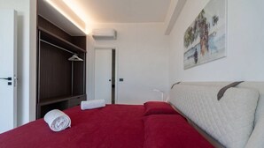 A hotel room with a red bedspread, a white headboard, a painting on the wall, and a wardrobe with a hanging rack. - Apartment Biancolilla - Maina Country Resort - with pool, in Sicily (Naso)