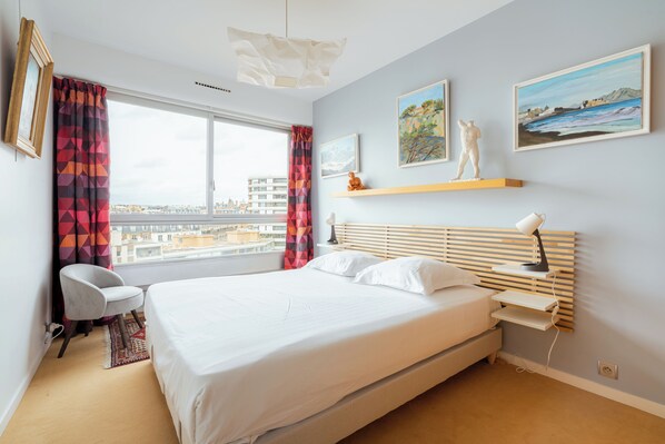 A modern bedroom with a large bed, a chair, a desk, and a view of the city. - The Salon in the Sky, by Veeve (Paris)