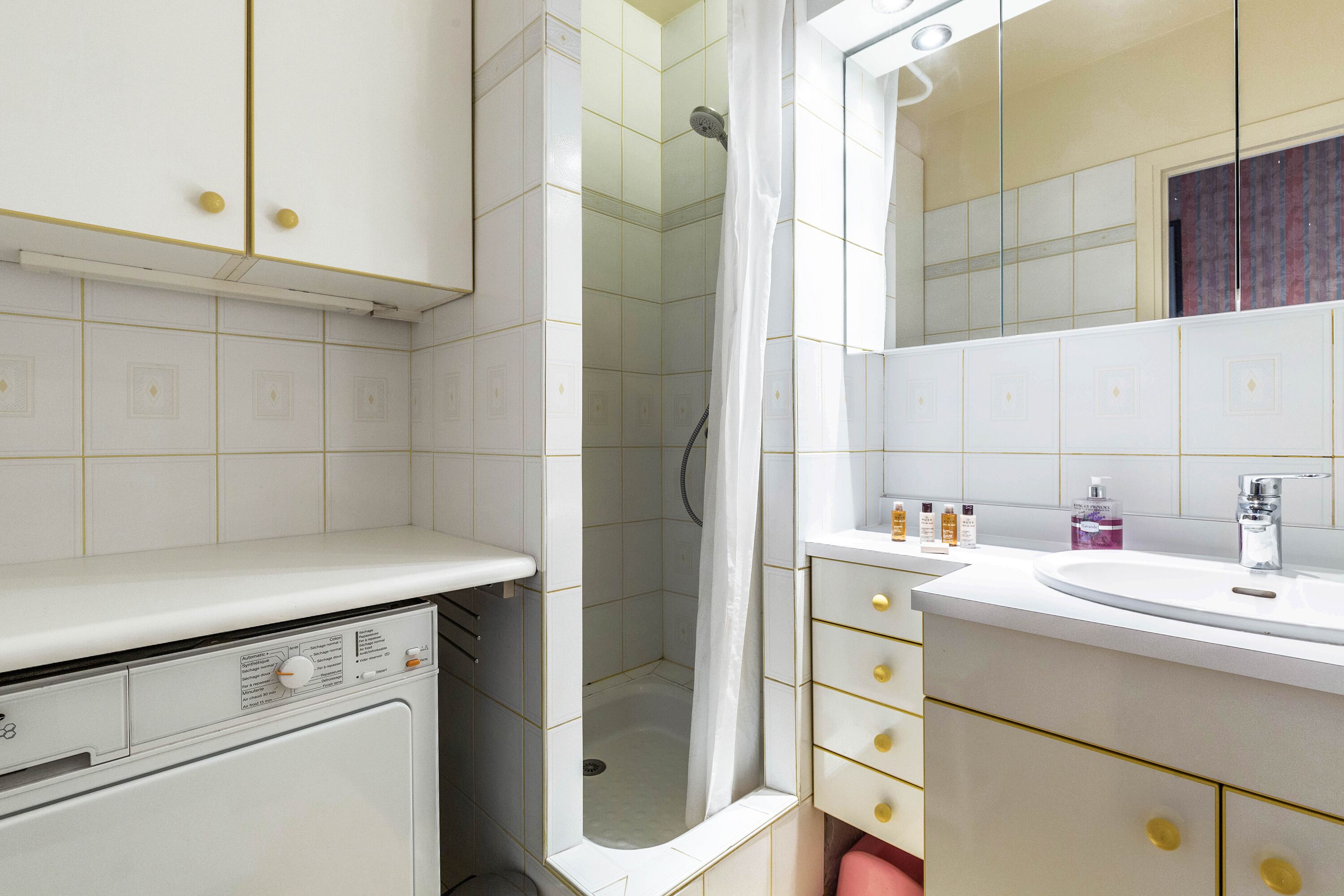 A bathroom with a white sink, mirror, and cabinet, a shower with a curtain, and a washing machine.