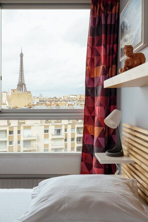 A hotel room with a bed, a desk with a lamp, a view of the Eiffel Tower, and a geometric-patterned curtain. - The Salon in the Sky, by Veeve (Paris)