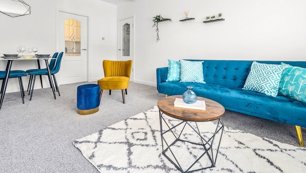 A modern living room with a blue sofa, yellow armchair, and a dining area with a table set for four.