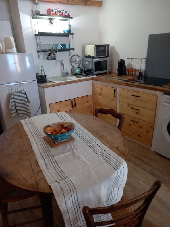 A compact kitchen with a dining table, chairs, a bowl of fruit, a washing machine, and a microwave.