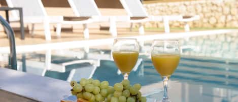 A poolside table with a plate of grapes and orange slices, two glasses of orange juice, and lounge chairs in the background.