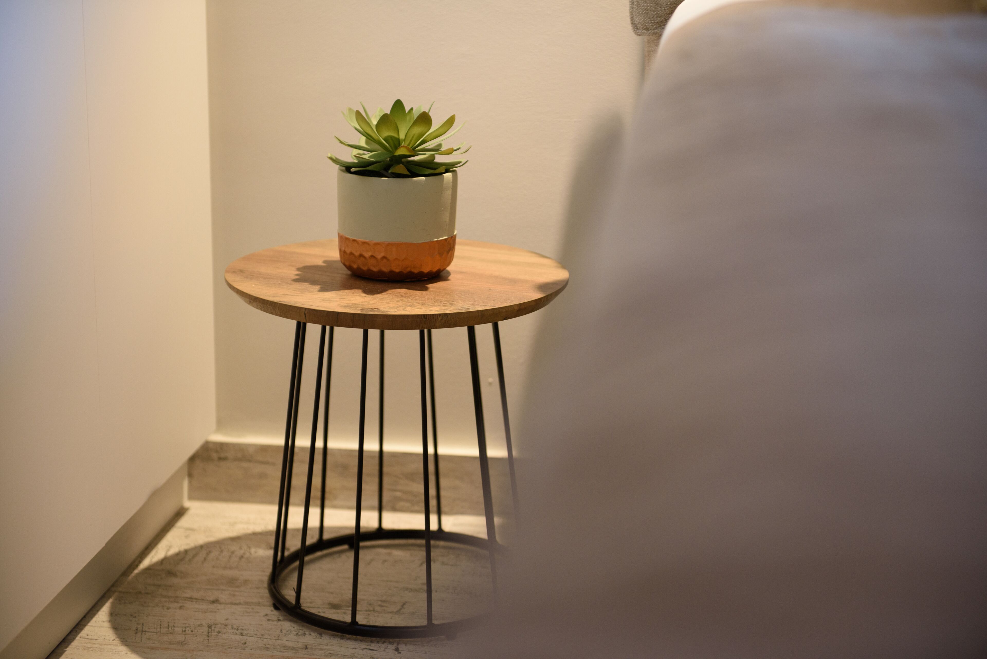 Un petit tabouret en bois, sur lequel est posée une plante en pot, est placé dans un coin d’une pièce dont le sol est de couleur claire et le mur est uni.