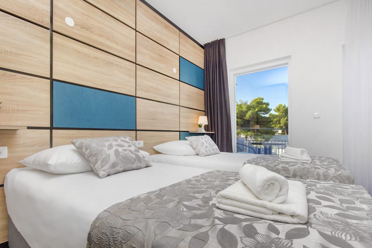 A hotel room with two beds, a wooden headboard, a view of trees through a window, and a balcony.