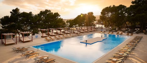 A pool area with sun loungers and cabanas, surrounded by trees and a clear sky.