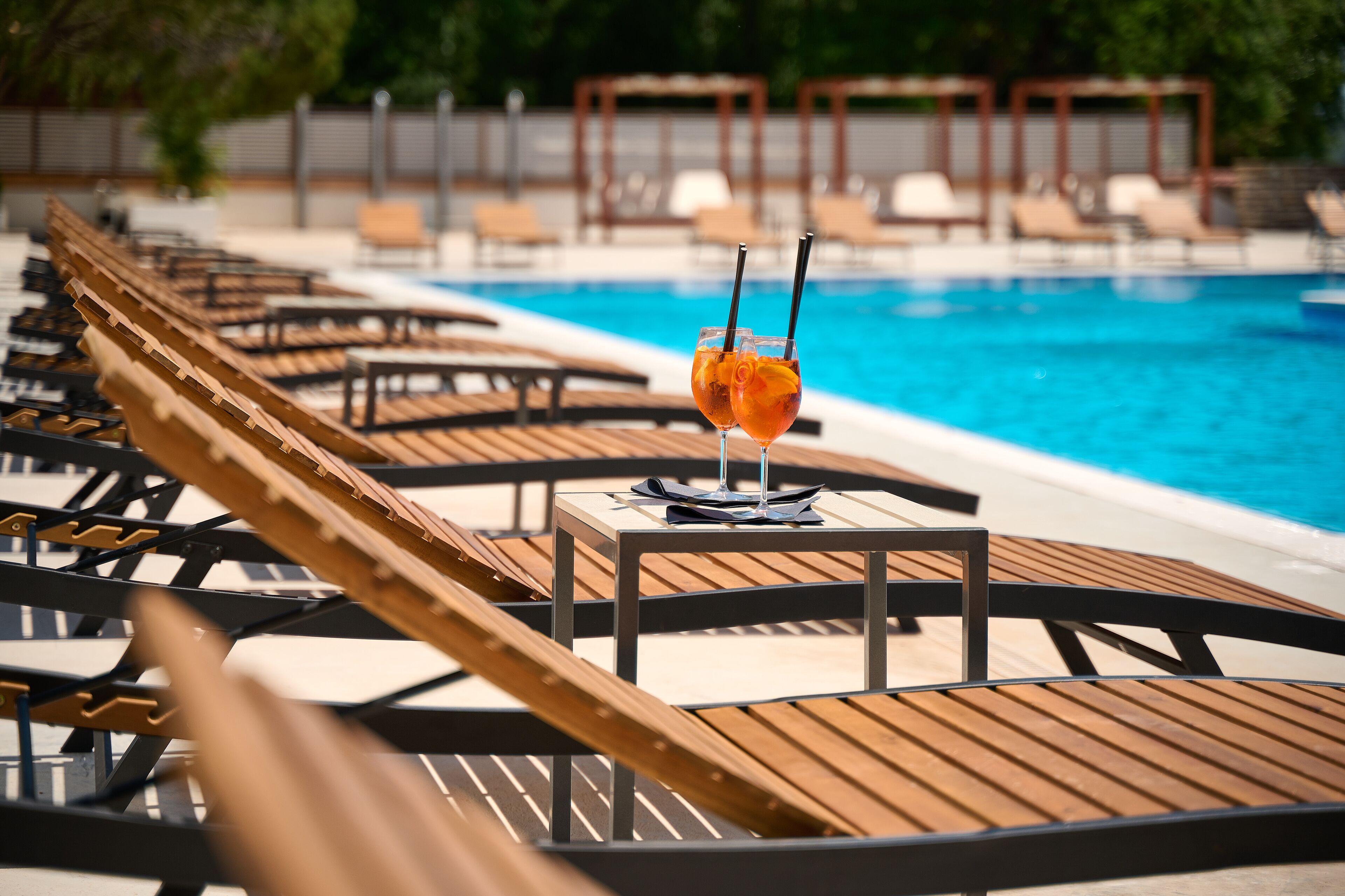 Un espace aménagé au bord de la piscine, avec des chaises longues en bois et une table sur laquelle est posée une boisson.