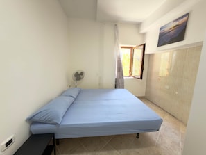 Una habitación pequeña y limpia con cama, ventilador, ventana con cortinas y un cuadro en la pared.
