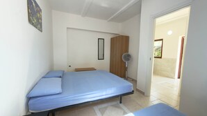 A bedroom with a bed, a wooden wardrobe, a standing fan, and a door leading to an outdoor area.