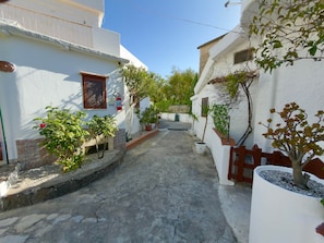 A narrow street with white buildings, potted plants, and a fire extinguisher mounted on a wall.