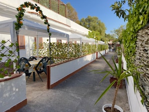 A covered outdoor area with a row of white buildings, a concrete floor, and potted plants.
